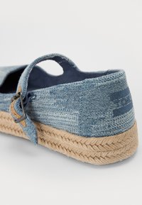 Sapatilha de denim de calçar, com padrão azul, faixa elástica e sola de juta trançada em tom de cáqui. Apresenta um recorte para respirabilidade e costura detalhada.