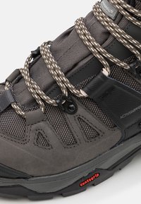 Grå vandringsko med texturerad tyg och läderdetaljer, utrustad med reflexsnören, ett säkert fästsystem och en hållbar sula.