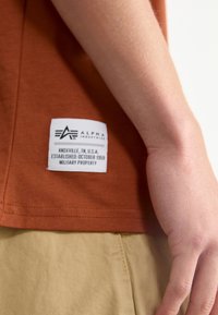 Tielko v rusty farbe s bielou nálepkou zobrazenou "Alpha Industries," založená 1959, a "Vojenský majetok." Hladká textúra látky.