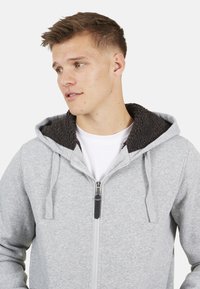 Grijze rits hoodie met donkere fleece voering in de capuchon. Beschikt over trekkoorden en een subtiele textuur. Draagt over een wit shirt.