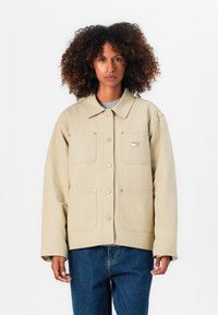 Beige jakke med kravehals, fire frontlommer, trykknaplukninger og et tekstureret stof, båret over en grå skjorte og blå jeans.