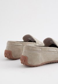 Beige suede slip-on shoes with a rubber sole and textured grip detail. Features stitched accents and padded openings at the heel.