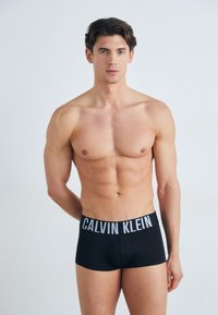 Juodi Calvin Klein bokseriai su pilku ženklintu juosmeniu. Medžiaga yra lygi, glaudžiai priglunda prie kūno ir turi švelnų tekstūrą.