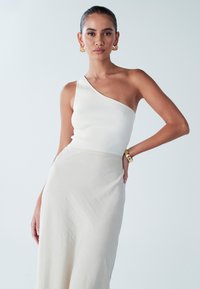 Haut côtelé à une épaule en blanc, associé à une jupe fluide crème. Le look est agrémenté de boucles d'oreilles dorées et d'un bracelet métallique. Texture lisse.