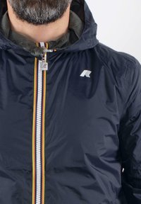 Giacca leggera navy con chiusura a zip, striscia multicolore, cappuccio e logo sul petto. Tessuto liscio e riflettente.