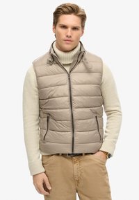 Padded vest w kolorze jasnego beżu, z zamkiem z przodu, wysokim kołnierzem, bocznymi kieszeniami i gładką, błyszczącą teksturą. Nosi się go na kremowym swetrze.