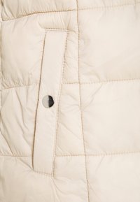 Detail béžové prošívané bundy s viditelným stříbrným drukem a klopovou kapsou s prošívanou texturou a šitými švy.