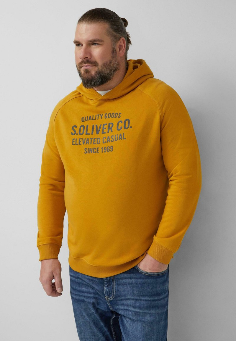 Mężczyzna z brodą w musztardowej bluzie z kapturem z napisem "S. Oliver Co. Elevated Casual Since 1969", w niebieskich dżinsach, stoi pod białą ścianą.