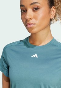 Camiseta deportiva de manga corta color verde azulado, confeccionada en una tela suave, con un logo blanco en el pecho y costuras planas para mayor comodidad.