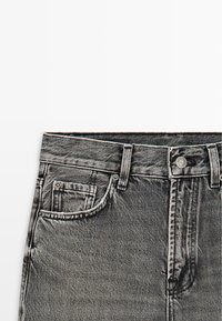 Jean en denim noir délavé montrant la ceinture, les passants de ceinture, le bouton en métal et la poche avant avec petite poche à monnaie sur fond blanc.