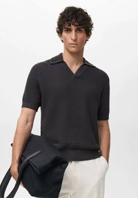 Polo shirt lavorato a maglia grigio scuro con maniche corte e scollatura a v. Il modello tiene in mano una borsa nera, dotata di manici rettangolari e una forma strutturata.