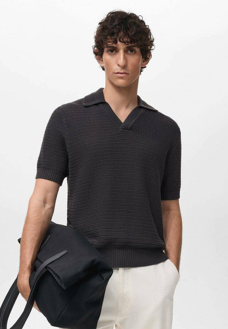 Polo shirt lavorato a maglia grigio scuro con maniche corte e scollatura a v. Il modello tiene in mano una borsa nera, dotata di manici rettangolari e una forma strutturata.