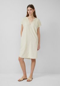 Hellgelbes Leinenkleid mit V-Ausschnitt, kurzen Ärmeln, lässigem Schnitt und knielangem Saum. Getragen mit geflochtenen, tanfarbenen Schlupfsandalen.