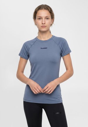 Ung kvinde med lys brun hår, iført en tætsiddende blå hummel t-shirt med korte ærmer og sorte hummel bukser, stående med hænderne foldet.