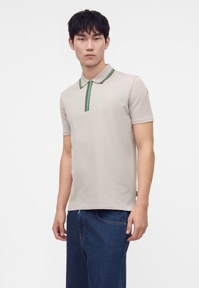 Jeune homme aux cheveux courts noirs portant un polo beige clair avec une bordure verte et un jean bleu foncé, debout devant un fond uni.