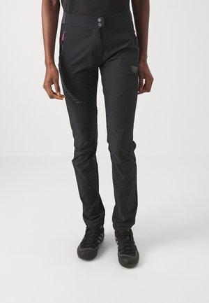 Person trägt eng anliegende schwarze Outdoorhosen mit Reißverschlusstaschen und schwarze Sportschuhe, steht vor einem einfarbigen Hintergrund.
