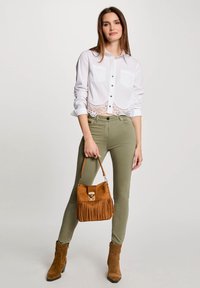 Weißes, kurzes, geknöpftes Hemd mit Spitzenbesatz, olivgrüne, enganliegende Hose, braune Wildleder-Stiefeletten und eine braune, fransige Handtasche.