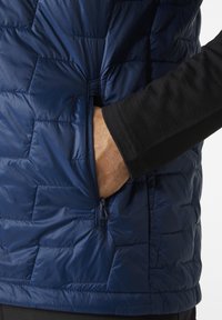 Chaleco acolchado azul marino con un diseño de costura, que presenta un bolsillo con cremallera y una capa de manga larga negra debajo. Textura suave y brillante.