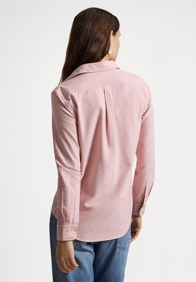 Svetloružová košeľa na gombíky s golierom, dlhými rukávmi a záhybom na chrbte. Tkanina pôsobí hladko s jemnou textúrou. Kombinovaná s modrými džínsami.