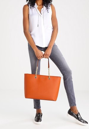 Sac fourre-tout orange avec texture en cuir, poignées en chaîne double et en cuir. Baskets rayées noir et blanc et jeans gris déchirés. Chemise blanche.