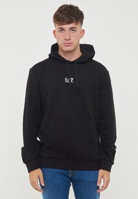 Svart hettegenser laget av myk stoff, som har en frontlomme som en kenguru, snor i hetten og en liten hvit logo på brystet.