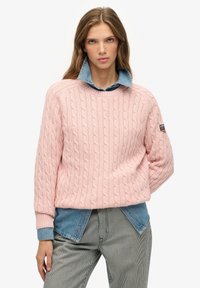 Jasnoróżowy sweter w warkocze z okrągłym dekoltem i ściśniętymi mankietami, nałożony na dżinsową koszulę z kołnierzykiem, w zestawie z spodniami w paski.