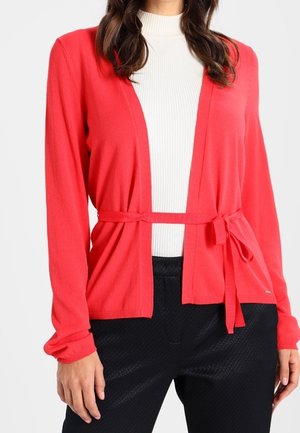 Frau trägt eine leuchtend rote, gebundene Strickjacke über einem weißen, gerippten Rollkragenpullover und schwarzen, strukturierten Hosen, dargestellt vom Hals bis zur Hüfte.