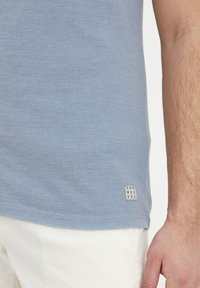 Svetlomodrá polokošeľa z textúrovanej tkaniny s malou, štvorcovou označovacou nálepkou na leme, skombinovaná s bielymi nohavicami.