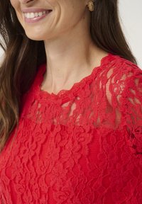 Haut en dentelle rouge avec un décolleté festonné et des motifs floraux délicats. Le tissu a un dessus transparent, accentuant l'apparence texturée.