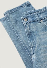 Světle modré džíny z denimu položené na rovné ploše, se zapínáním na knoflík, prošitými okraji a mírně vybledlým vzhledem, obsahují boční kapsy.