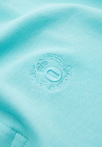 Tyrkysová látka s texturovanou tkaninou, která obsahuje vyšívané logo ve tvaru kruhu obklopeného vavřínovými listy.