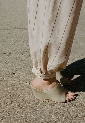 Personne portant un pantalon beige à fines rayures retroussé à la cheville et des mules beiges à talon compensé et bout ouvert, debout sur un sol texturé.