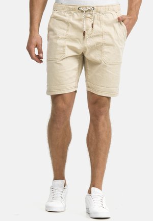 Mand iført beige shorts med snøre, frontlommer og hvide sneakers, stående mod en ensfarvet baggrund.