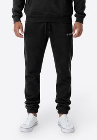 Zwarte joggingbroek van zacht materiaal, met een elastische tailleband met trekkoord en elastische boorden bij de enkels. Minimale branding zichtbaar.