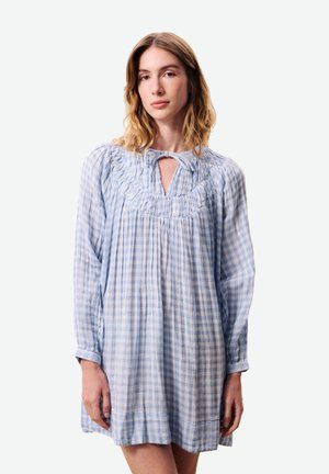 Jeune femme aux cheveux blonds mi-longs portant une robe à manches longues à carreaux bleu clair et blanc avec un décolleté en trou de serrure.