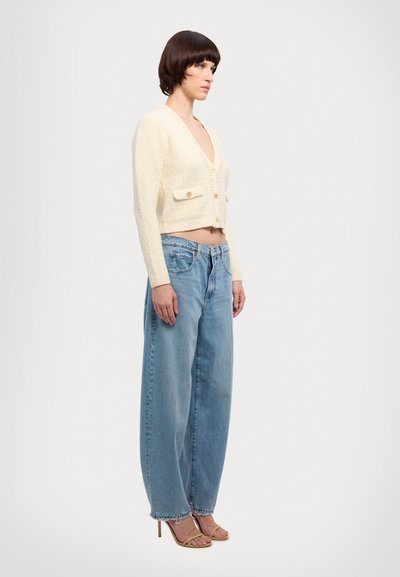 Femme portant un cardigan crème boutonné, un jean ample bleu clair et des sandales beige à talons hauts, debout de profil sur fond blanc.