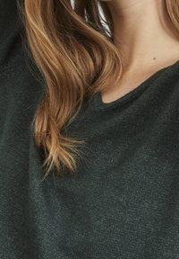 Maglione a maglia verde scuro con scollatura a V, che mostra una texture morbida e delle sfumature sottili, indossato da una persona con capelli lunghi e mossi.