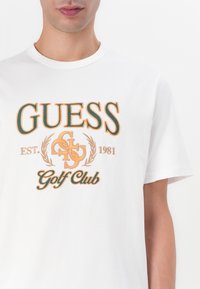 Camiseta blanca de algodón con texto bordado en naranja y verde en negrita: "GUESS EST. 1981 Golf Club," con acentos decorativos de laureles.