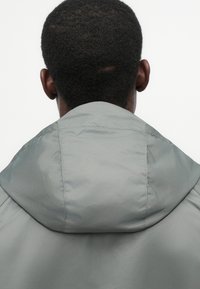 Ανοιχτό γκρι μπουφάν με κουκούλα, με λεία, ματ υφή και εμφανείς λεπτομέρειες ραφής, βλέπεται από την πίσω πλευρά.