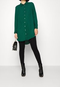 Groen overhemd met lange mouwen en knopen, voorzien van een kraag, gecombineerd met zwarte legging en enkellaarzen. Glad stof, afgeronde zoom en zilveren knopen.