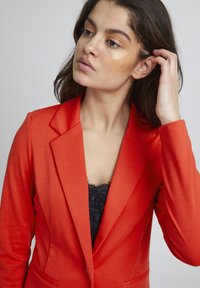 Rode, getailleerde blazer met scherpe revers en twee voorkruiszakken. Draag het over een zwarte kanten top, die een gladde textuur en een op maat gemaakt ontwerp laat zien.