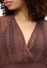 Κοντινό πλάνο μιας γυναίκας που φοράει καφέ δαντελωτό τοπ με V-λαιμόκοψη και περίπλοκα λουλουδένια και γεωμετρικά σχέδια, με εμφανή τα σγουρά μαλλιά της.