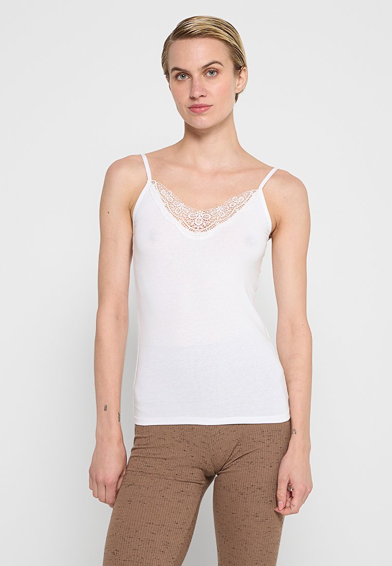 Weißes Top mit dünnen Trägern, das mit einem Spitzenbesatz am Hals ausgeschnitten ist. Weicher Stoff mit einer taillierten Form. Kombiniert mit braunen strukturierten Leggings.