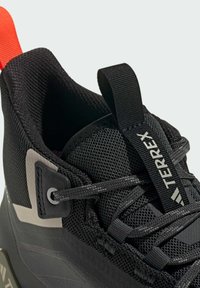 Sorte mesh atletiske sko med en tekstureret overflade, grå og sorte accenter samt en orange trækflig. Har et logo på siden og snøresystem.