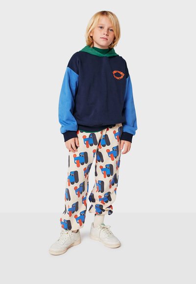 Felpa color block blu navy e blu con logo arancione, abbinata a pantaloni crema con motivi di cartoni animati colorati, indossata con sneakers bianche.