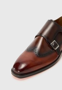 Zapato de cuero marrón con una forma elegante. Presenta una correa texturizada y una hebilla plateada. Perforaciones decorativas adornan la zona de los dedos.