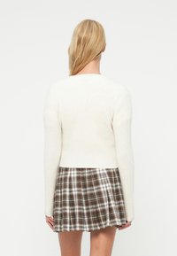 Λευκό πλεκτό πουλόβερ με μακριά μανίκια και στρογγυλή λαιμόκοψη, συνδυασμένο με καφέ πλισέ καρό φούστα με λευκές και μπεζ λεπτομέρειες.