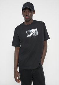 Μαύρο βαμβακερό μπλουζάκι με κοντά μανίκια, με μονόχρωμο γραφικό σχέδιο παπουτσιού στο μπροστινό μέρος. Συνοδεύεται από ένα ταιριαστό μαύρο καπέλο.