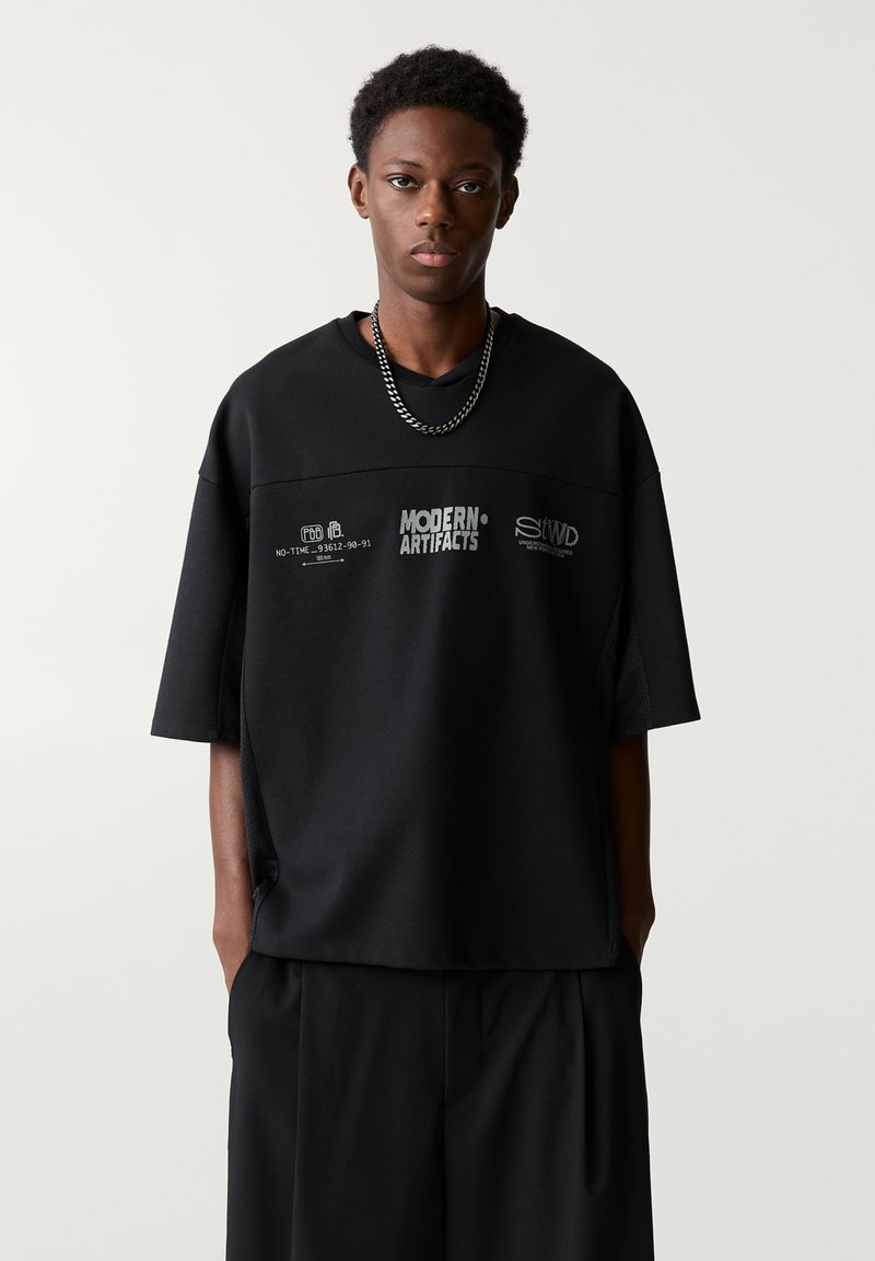 T-shirt nera oversize con maniche corte, caratterizzata da loghi e testi stampati. Tessuto morbido con una vestibilità ampia, abbinata a pantaloni neri.