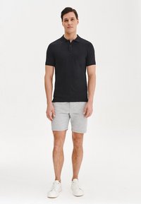 Czarna koszulka polo z krótkim rękawem, zestawiona z jasnoszarymi szortami. Model ma na sobie białe trampki; gładki materiał z minimalnym zdobieniem.